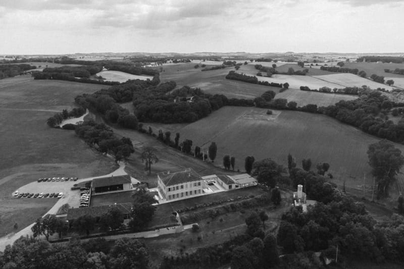 Black and white aerial drone shot of French country estate with manor house, outbuildings, and surrounding fields