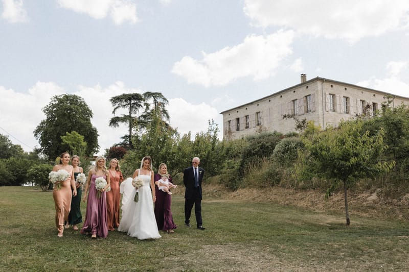 Bride and five bridesmaids in mismatched green terracotta mauve and plum dresses walking across manor lawn