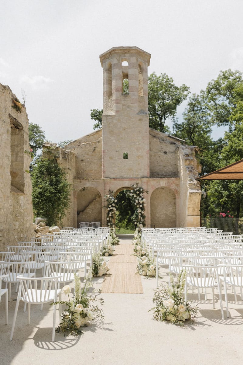 Ceremony in roofless stone chapel ruins with white floral arch and ivory aisle arrangements