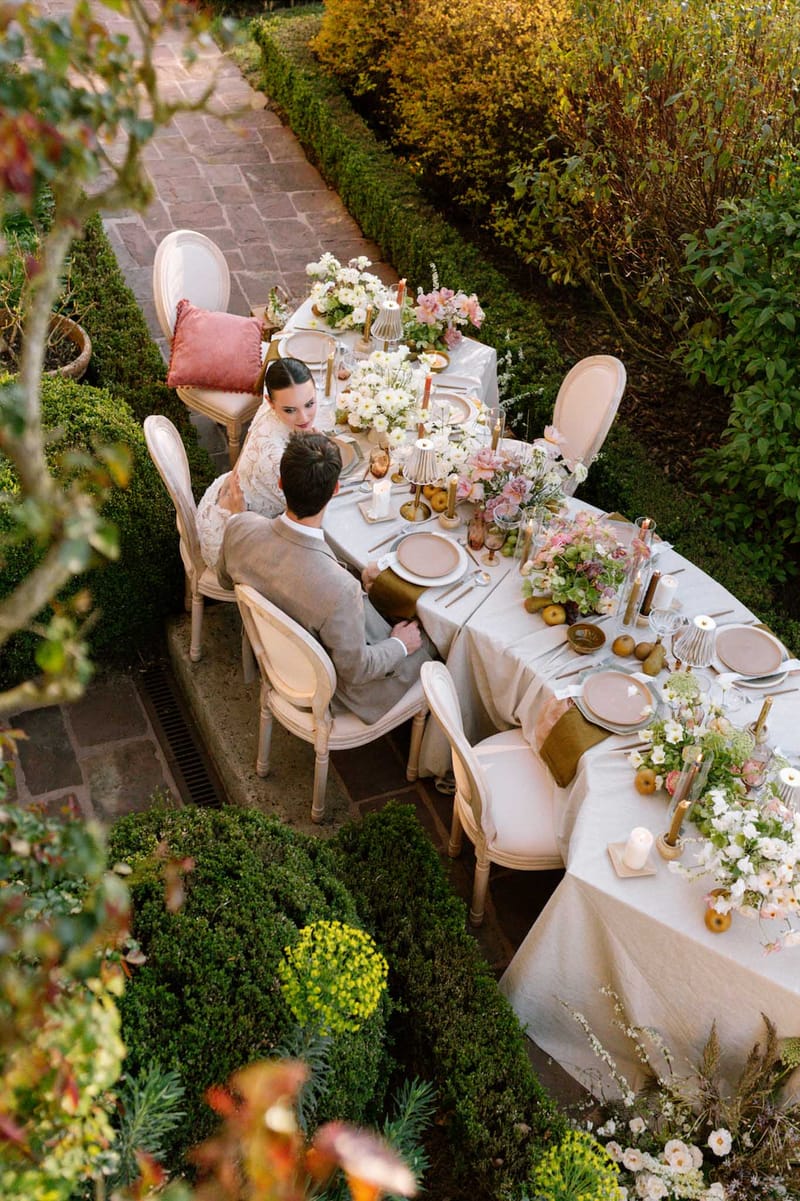 Aerial view of couple at an intimate garden reception table with blush and ivory florals, gold accents, and golden pears