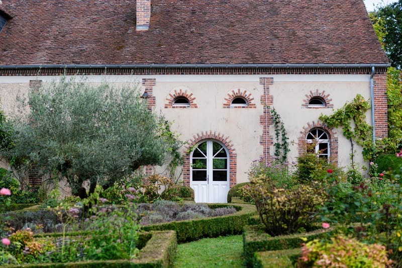 French country manor facade with cream walls, red brick arches, and formal parterre garden with boxwood hedges