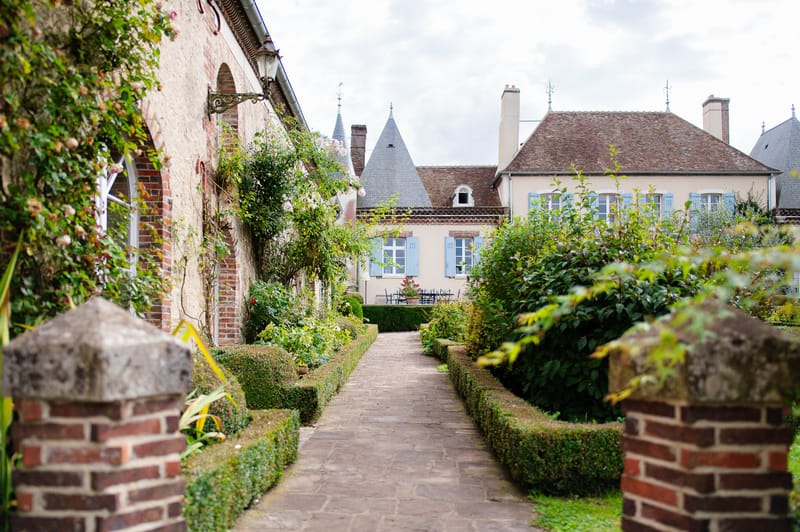 French provincial garden pathway with boxwood hedges leading to cream buildings with powder blue shutters