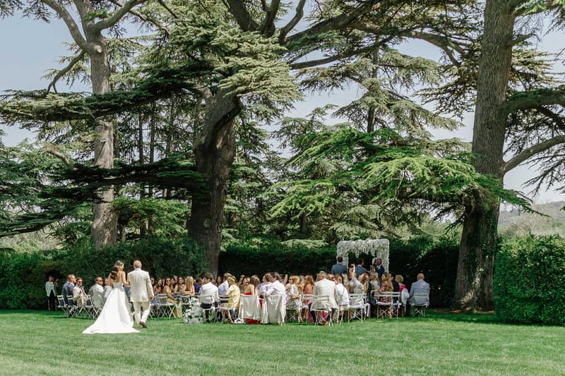Bride and groom walking down the aisle toward a white floral arch beneath cedar trees with seated guests on a lawn