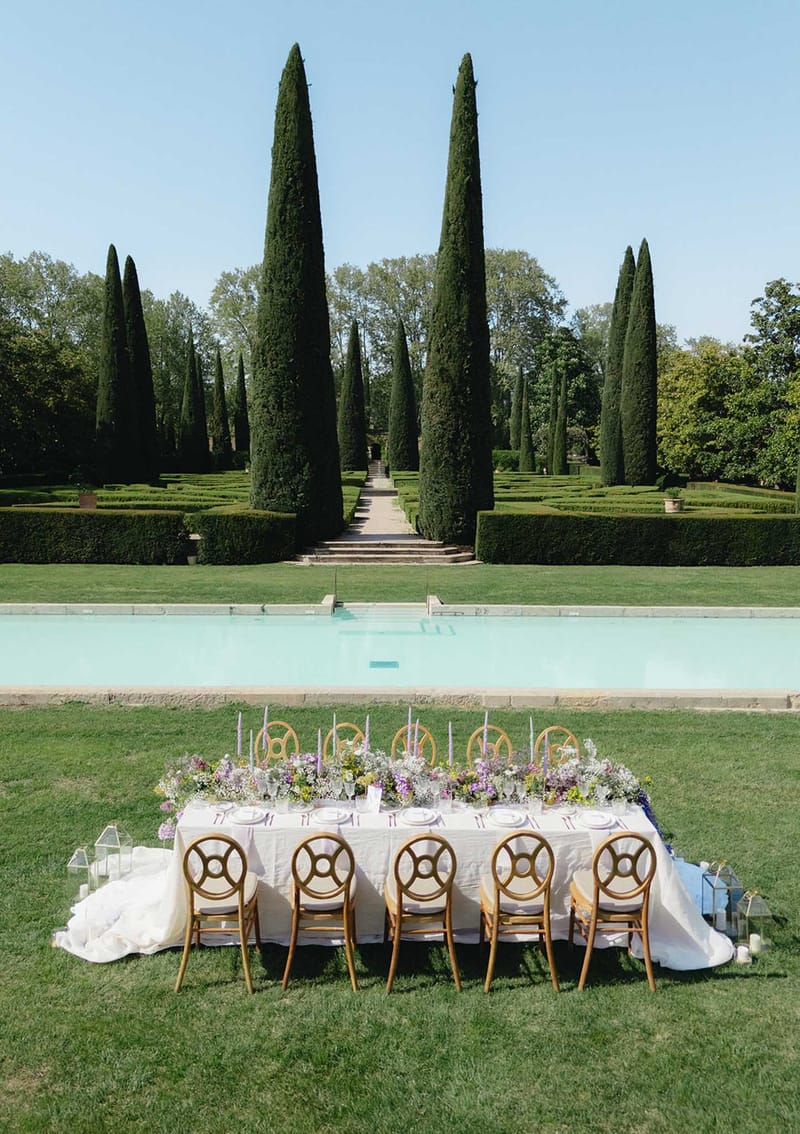 Formal garden reception table with purple and lavender flowers beside reflecting pool and cypress trees