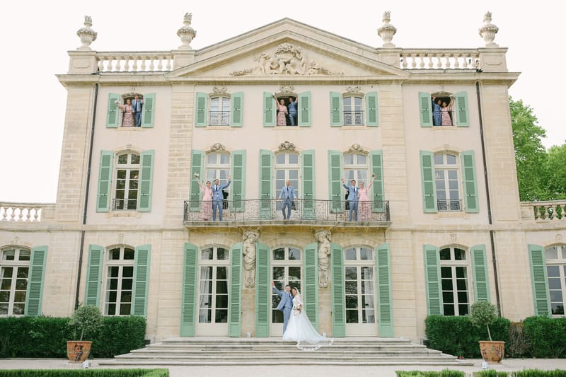 Bridal party posed across three levels of pink chateau facade with bride and groom on steps and attendants at windows