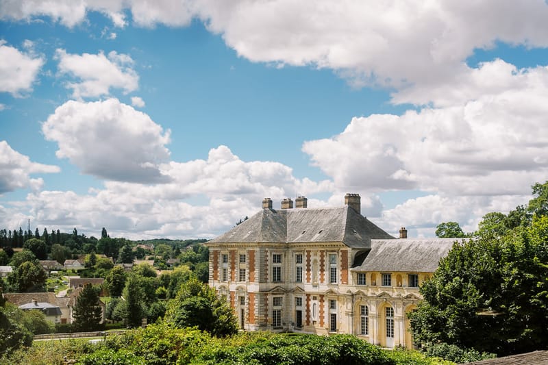 Classical French chateau with red brick, slate mansard roof, and dormer windows amid mature trees
