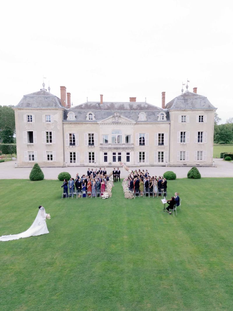 Aerial bride processing across lawn toward floral arch with sixty seated guests before classical French chateau