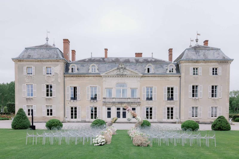Outdoor ceremony with ghost chairs and blush floral arch in front of classical French chateau