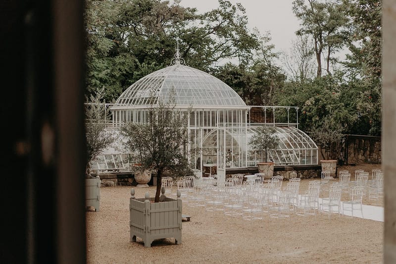 Ceremony setup with ghost chairs and white aisle runner facing Victorian glass greenhouse