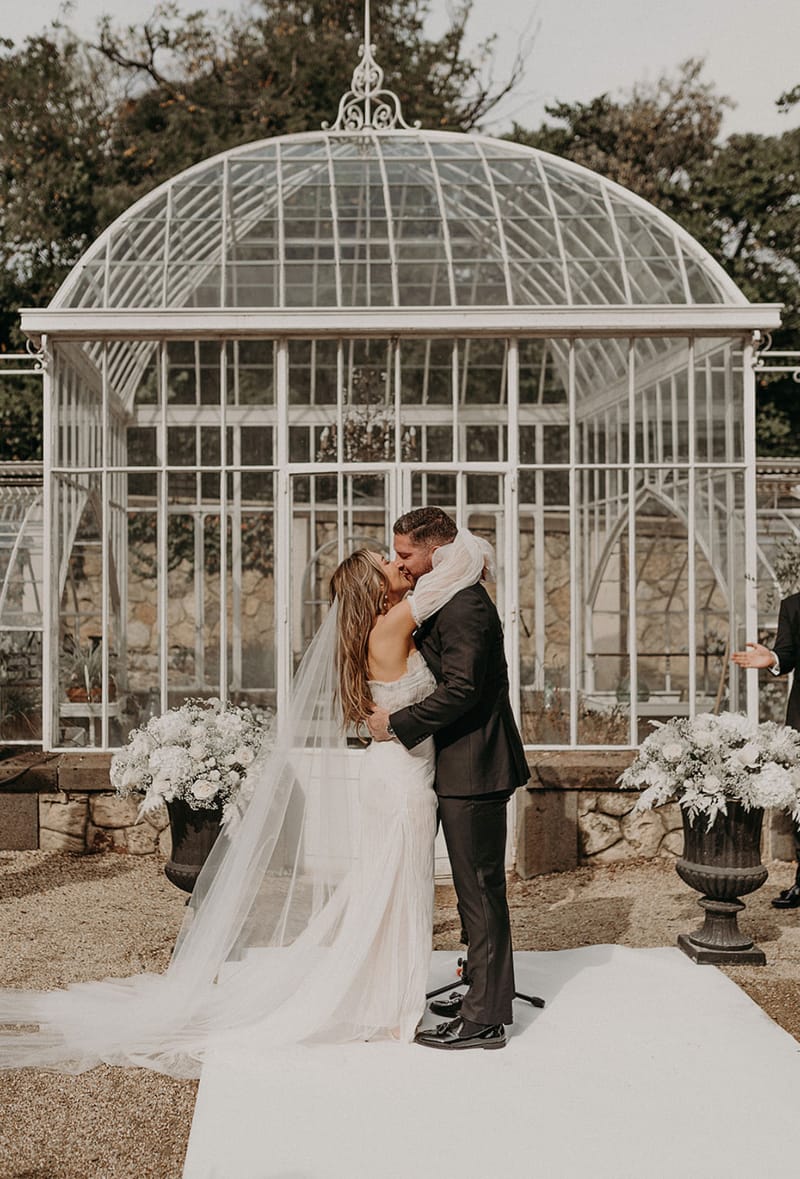 First kiss in front of Victorian glass greenhouse with white hydrangea urns