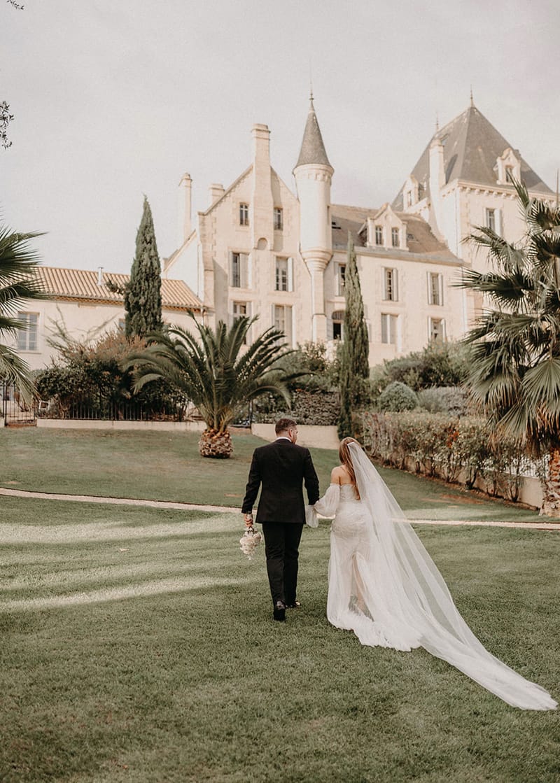 Couple walking toward turreted chateau from behind bride in off-shoulder lace gown with cathedral veil
