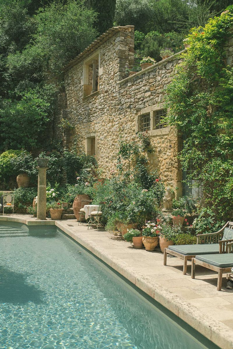 Rectangular pool with green-cushioned loungers and terracotta pots of pink geraniums beside stone mas