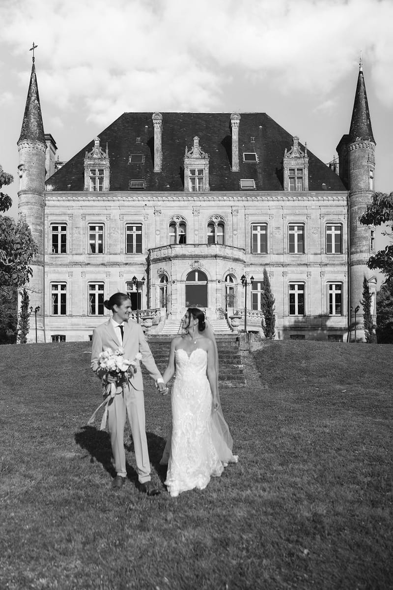 Couple walks hand-in-hand before Renaissance chateau with conical towers holding bouquet in B&W