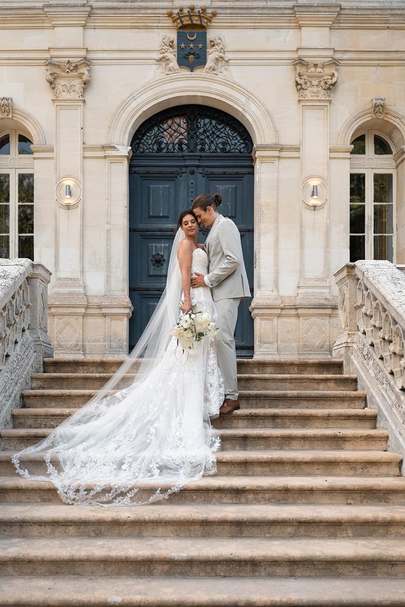 Bride and groom on chateau staircase with lace veil cascading down steps before teal arched door