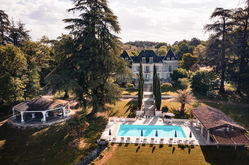 Aerial view of stone chateau with turquoise pool, white gazebo, and cypress-flanked grand staircase