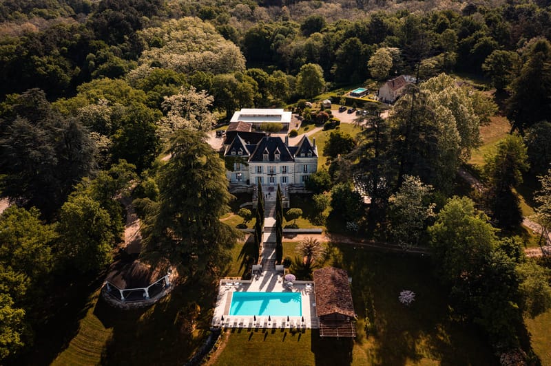 Aerial drone view of French chateau estate with mansard roofs, cypress-lined staircase, and turquoise swimming pool
