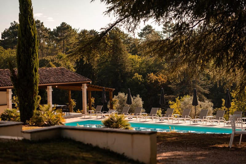 Turquoise pool with loungers and charcoal umbrellas beside stone-columned pergola at golden hour