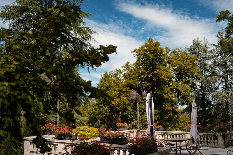 Stone terrace with balustrade, red flowering planters, iron lamp posts, and bistro seating at a chateau