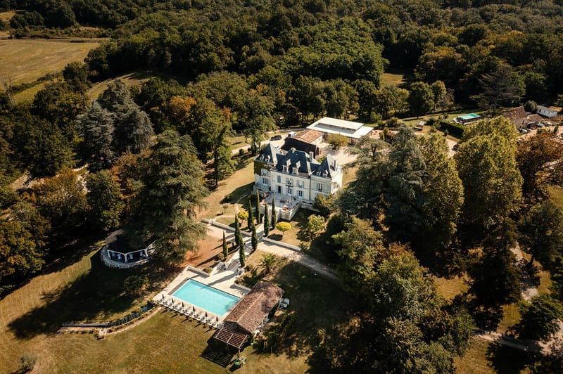 Aerial view of white French chateau with slate roof, swimming pool, event marquee, and surrounding woodland