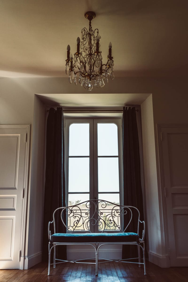 Symmetrical chateau room with wrought-iron bench, teal cushion, arched French doors, and brass chandelier