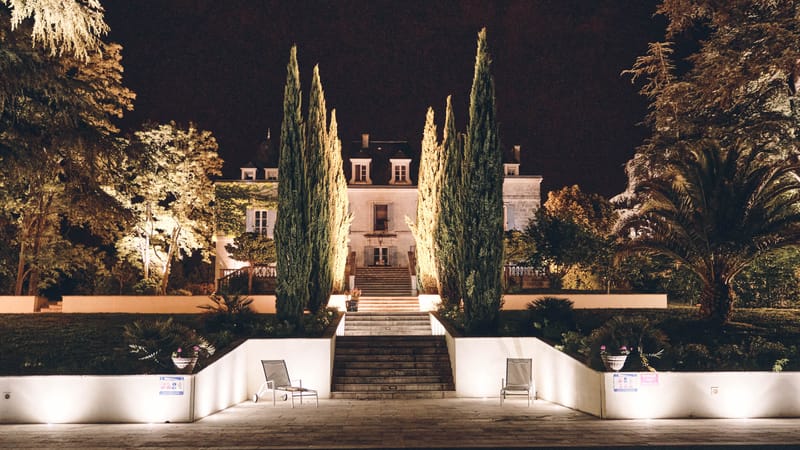 French chateau exterior at night with amber uplighting, cypress trees, stone staircase and pool terrace