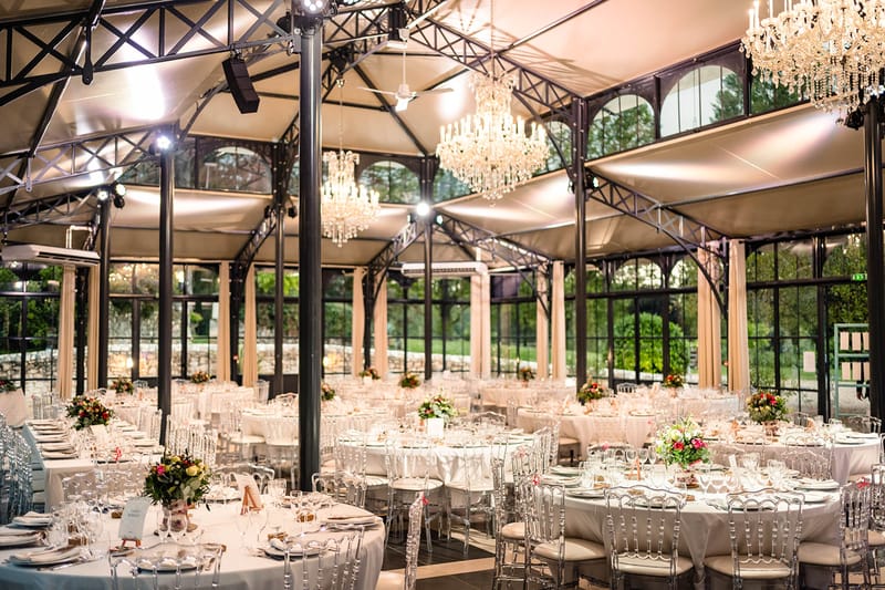Greenhouse wedding reception with crystal chandeliers, round tables, ghost chairs, and colorful floral centerpieces