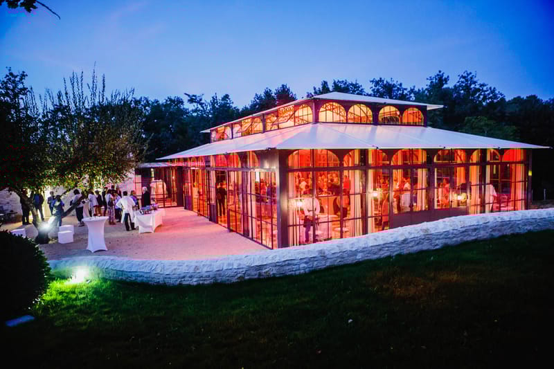 Evening wide-angle view of glass orangery venue with pink uplighting and guests on outdoor terrace at dusk