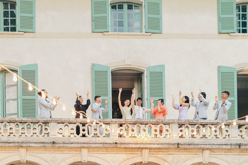 Bride in sequined dress and groom celebrate with guests on chateau balcony with string lights at night