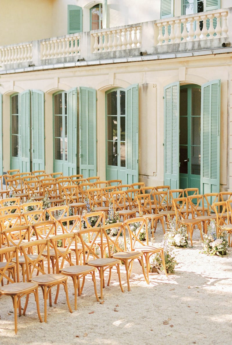 Ceremony setup with cross-back chairs and blush blue aisle florals before sage-shuttered chateau