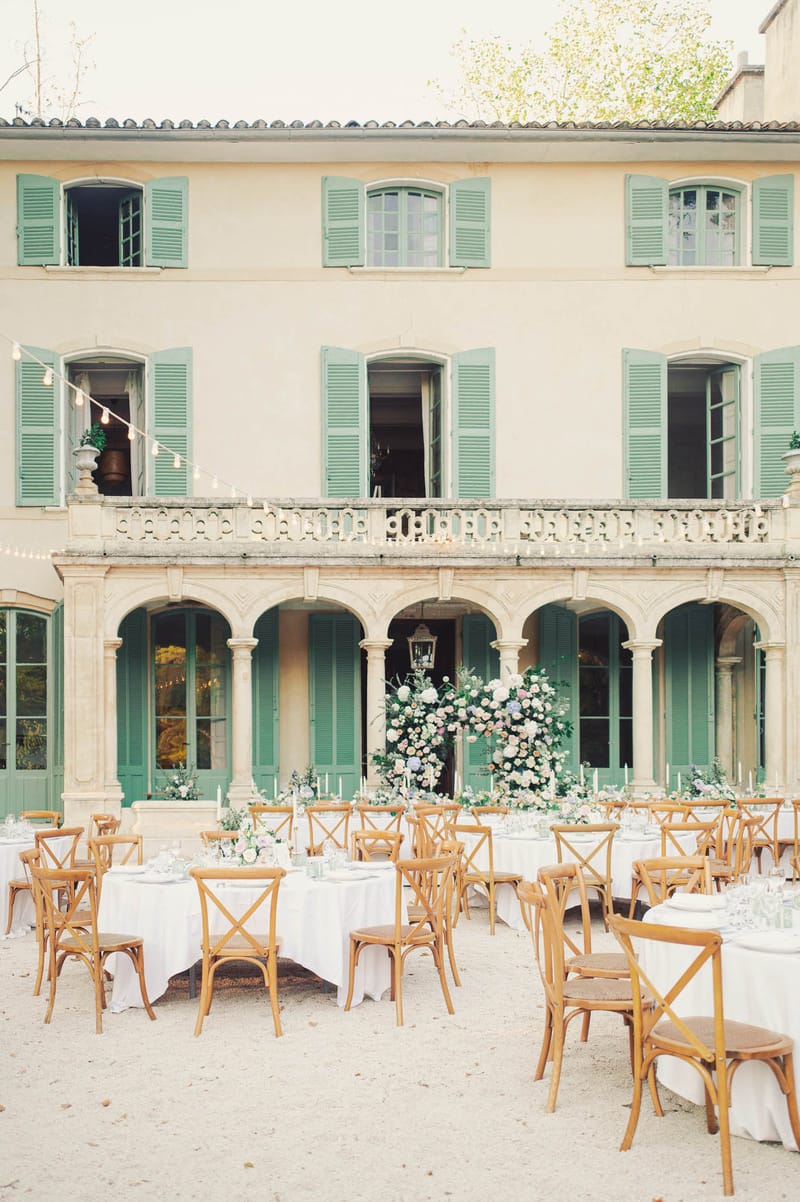 Outdoor courtyard reception with round tables, cross-back chairs, pastel floral centerpieces, and French manor facade with...