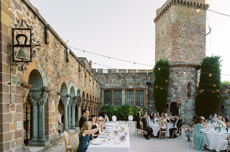Guests toasting at long tables in medieval courtyard with stone tower, lavender florals, and bistro string lights