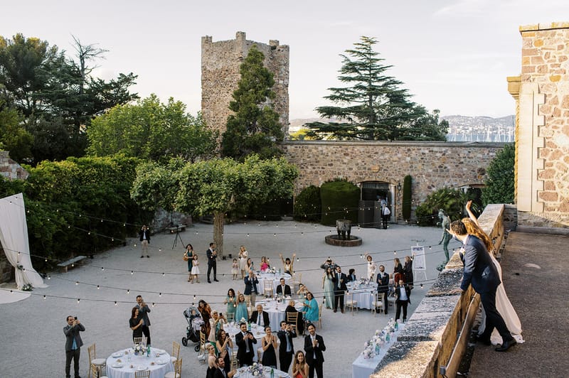 Couple waving from stone terrace above courtyard reception with round tables festoon lights and medieval tower behind