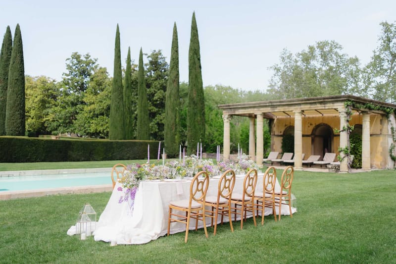 Long white table with lavender and wildflower runner and purple taper candles beside pool and stone colonnaded pavilion