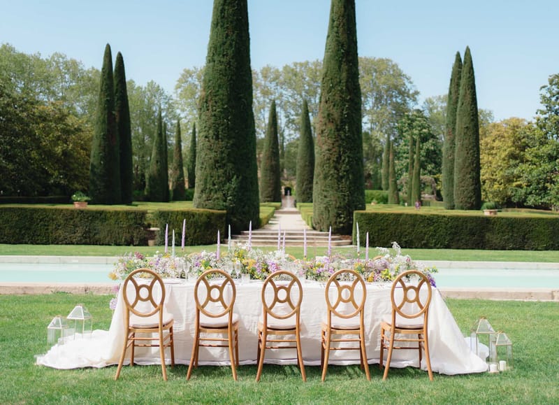 Reception table with ivory linen, lavender taper candles, and wildflower runner set before formal French garden with refle...