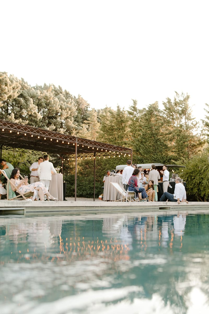 Golden hour cocktail reception beside pool with festoon-lit pergola, mobile bar, and guests reflected in still water