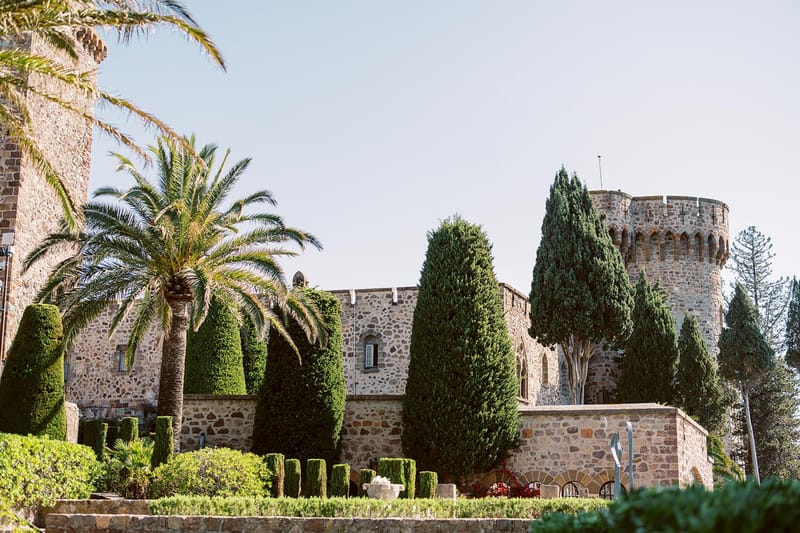 Stone castle wedding venue with rounded tower, battlements, topiary hedges, cypress trees, and palm tree in bright daylight