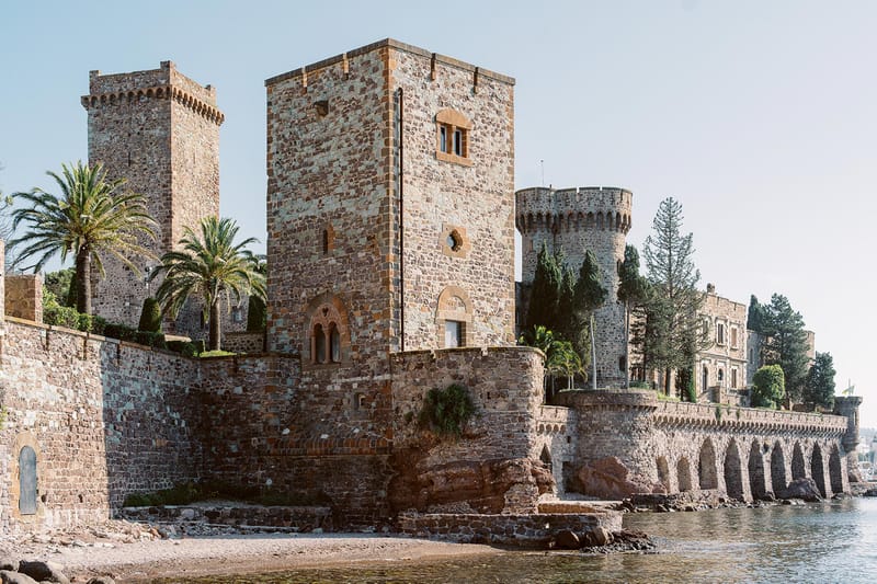 Medieval waterfront castle with crenellated towers, arched sea wall, and palm trees on rocky shoreline
