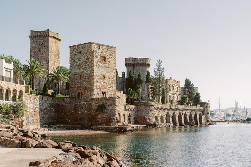 Medieval stone castle with crenellated towers and arched colonnades along the waterfront with palm trees
