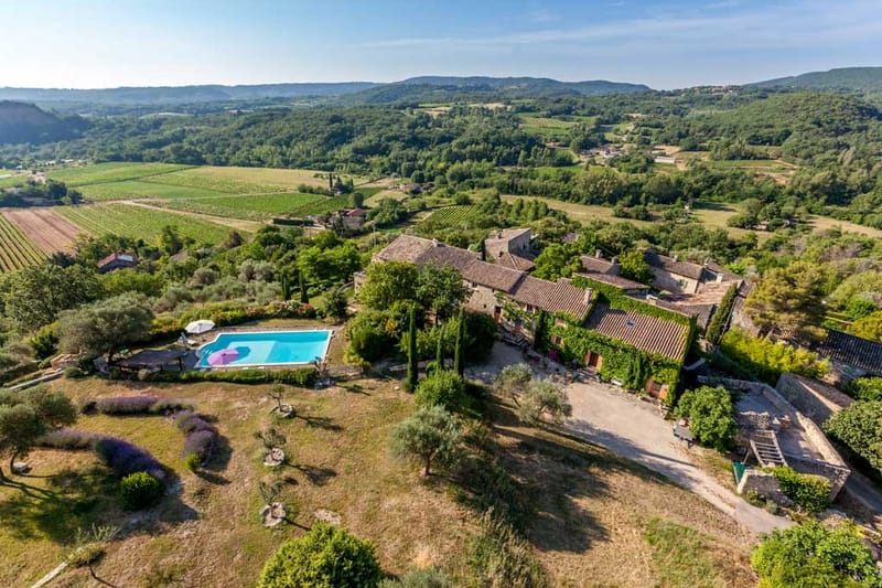 Aerial view of stone farmhouse complex with pool, lavender, and vineyard rows in rolling landscape