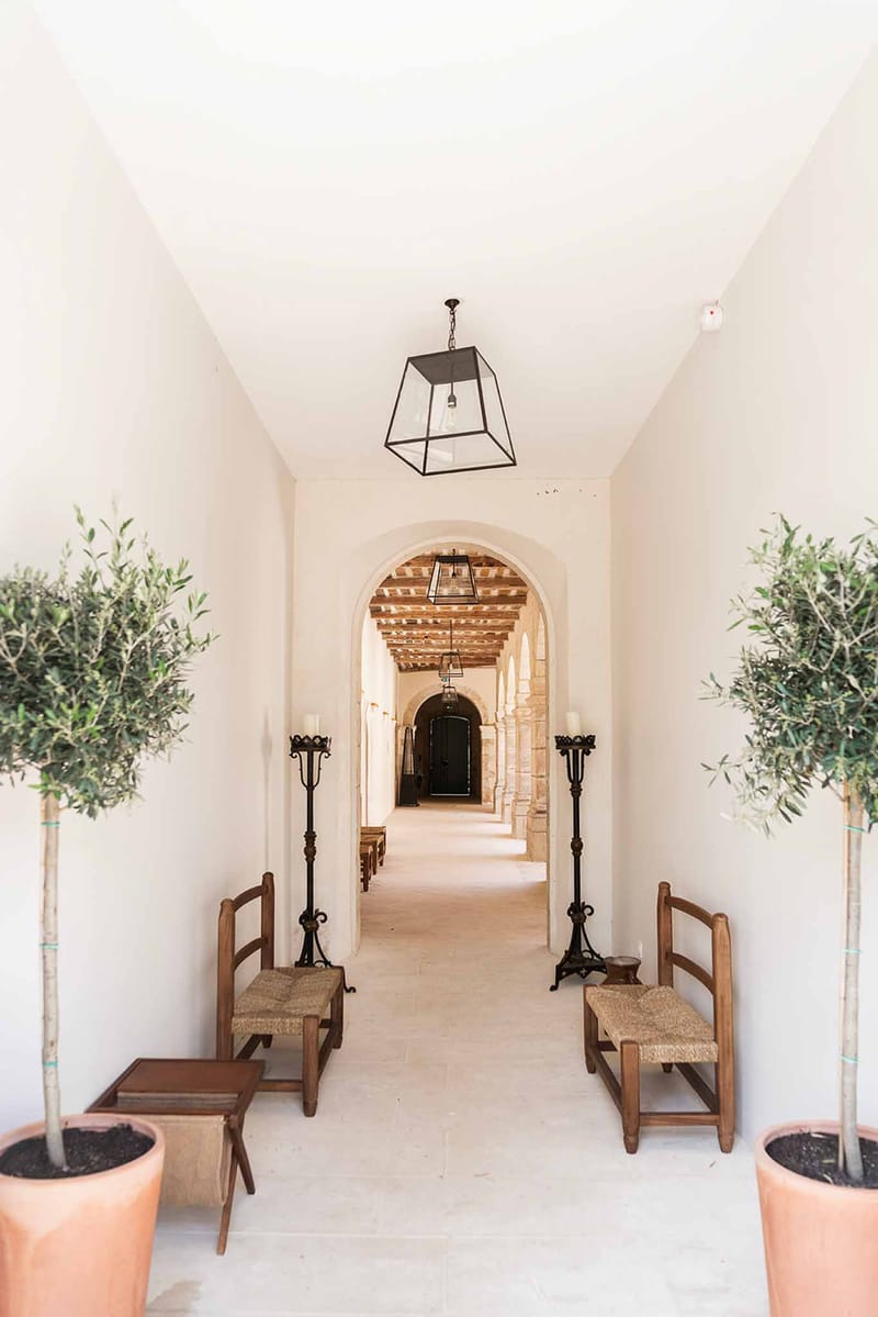 Chapel corridor with arched doorways, iron pendant lanterns, pillar candles, and potted olive trees