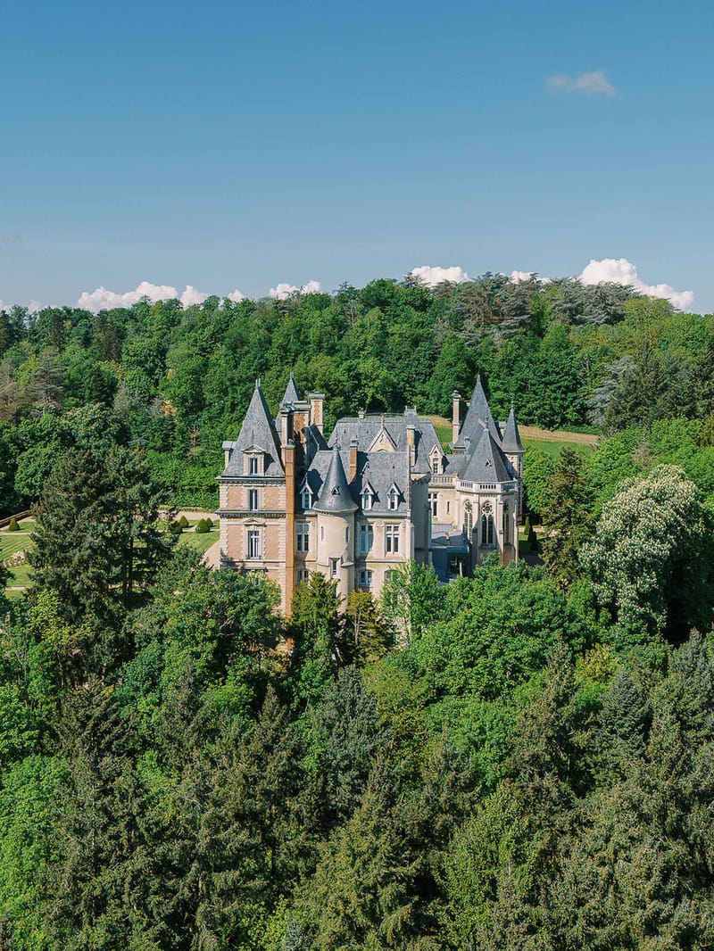 Aerial drone view of a French chateau with slate-grey turrets surrounded by mature trees and formal gardens