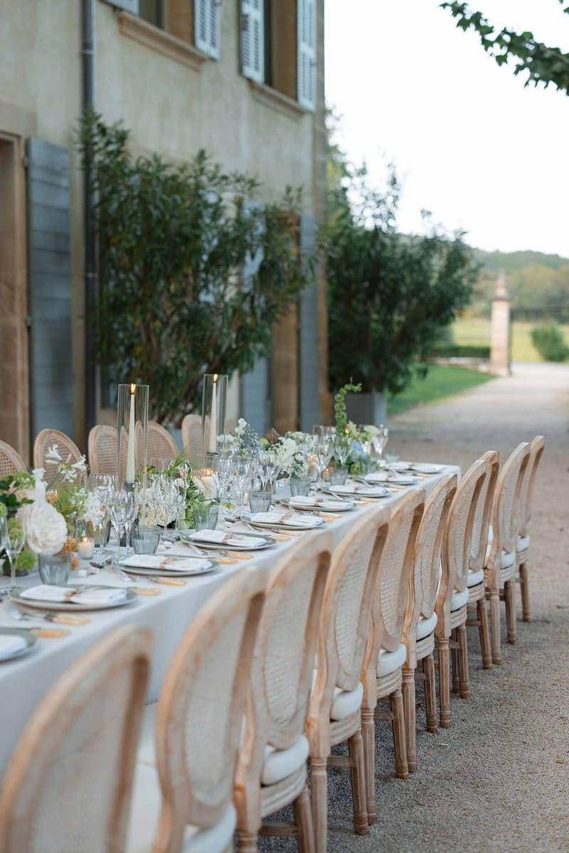 Long grey linen table with white floral centerpieces and cane-back Louis XVI chairs set before chateau facade