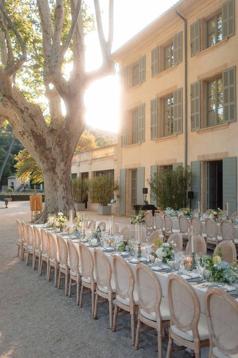 Long outdoor reception table with blue hydrangea centerpieces and gold candles at golden hour in Provencal courtyard