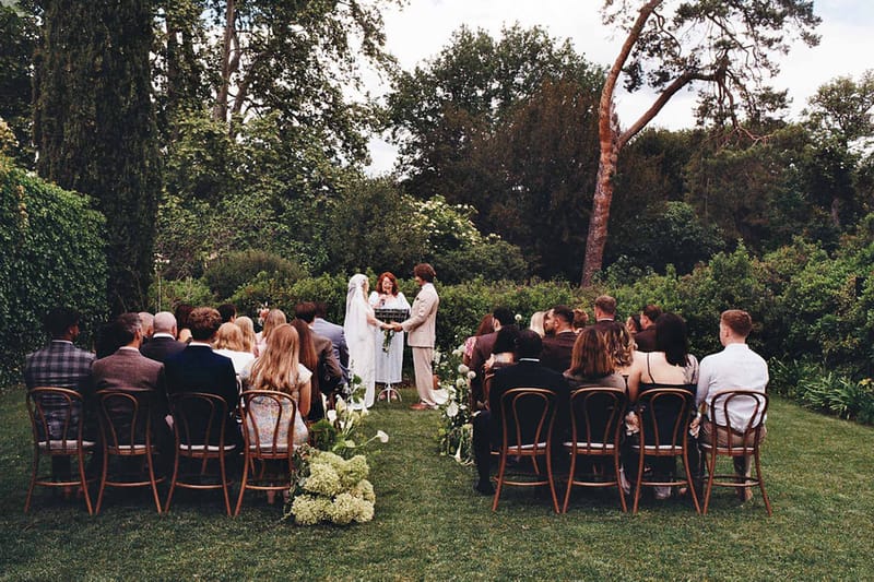 Thirty guests on bentwood chairs watch couple exchange vows on garden lawn with white hydrangea aisle flowers
