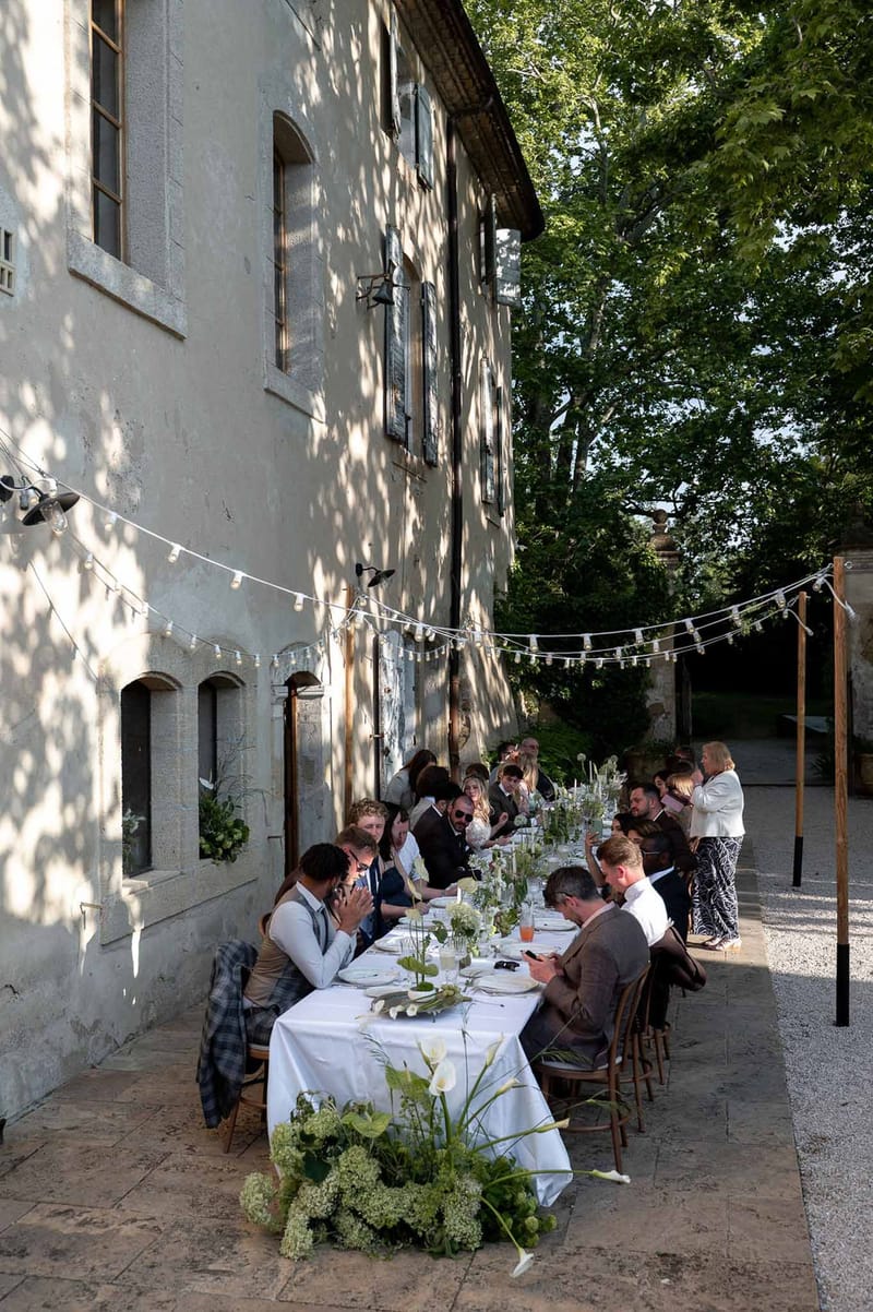 Twenty-five guests at banquet table with calla lily runner bistro lights and bentwood chairs along manor facade
