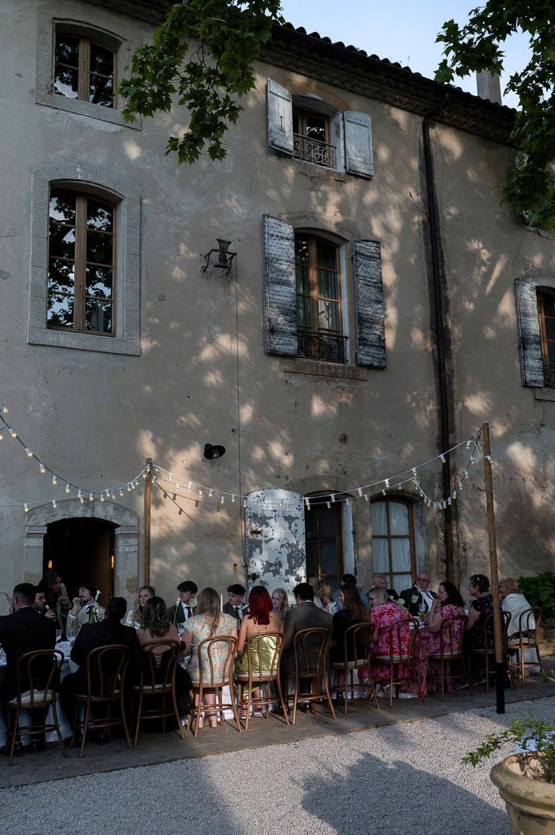 Guests at long tables along aged bastide facade with festoon lights, bentwood chairs, and colourful attire at dusk