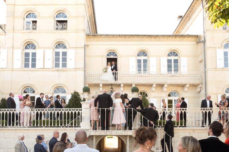 Couple on chateau balcony overlooking 30 guests gathered on terrace during cocktail hour in late afternoon light