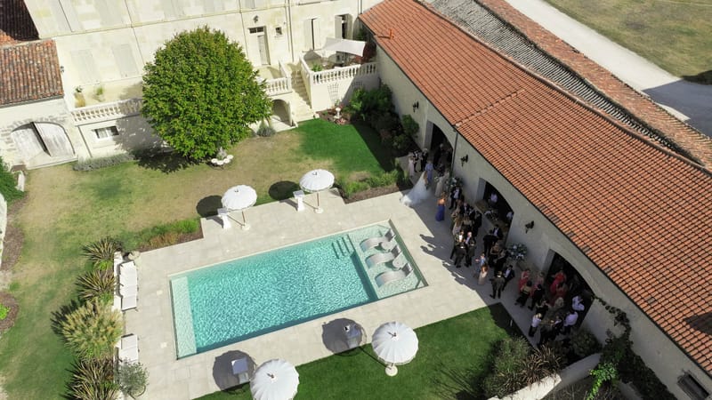 Aerial view of chateau with stone arcade, pool terrace, and 40 guests at cocktail hour in courtyard