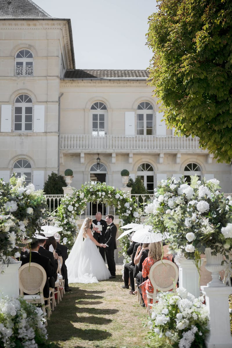 Ceremony with circular white floral arch and pedestal arrangements before limestone chateau facade