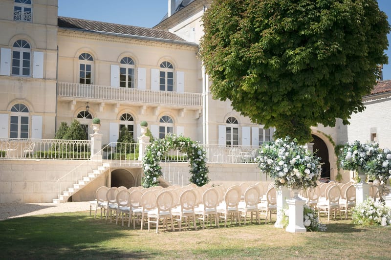 Ceremony arch of white roses and greenery with rattan chairs before two-storey limestone chateau
